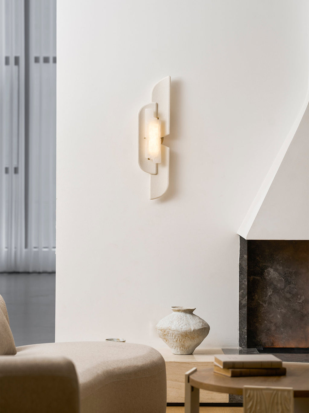 A Clérachic wall lamp hangs high on a white plaster wall beside a sheer curtain, over a cream chair and a fireplace.