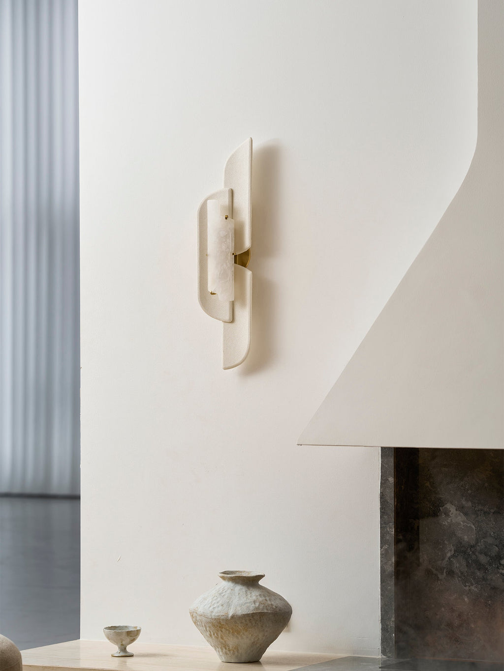 A Clérachic wall lamp on a white curved wall above a small ceramic vase and a stone hearth, seen from a lower angle.