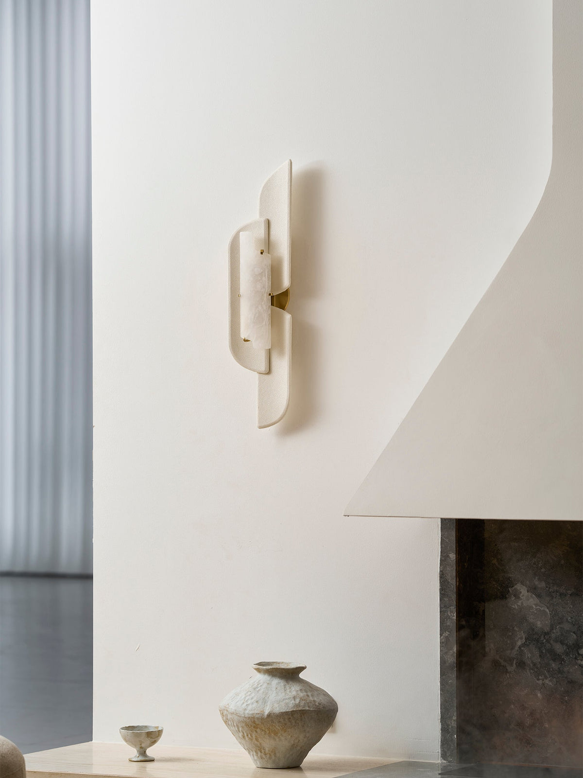 A Clérachic wall lamp on a white curved wall above a small ceramic vase and a stone hearth, seen from a lower angle.