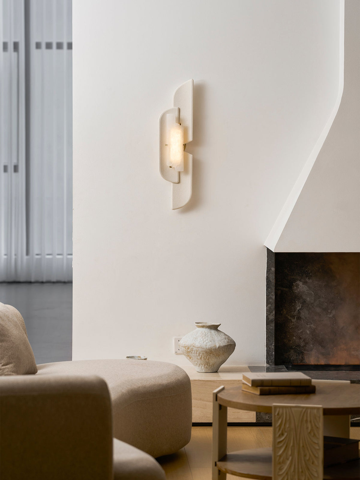 A lit Clérachic wall lamp above a cream sofa and a round wood table, beside a stone fireplace and a pale vase.