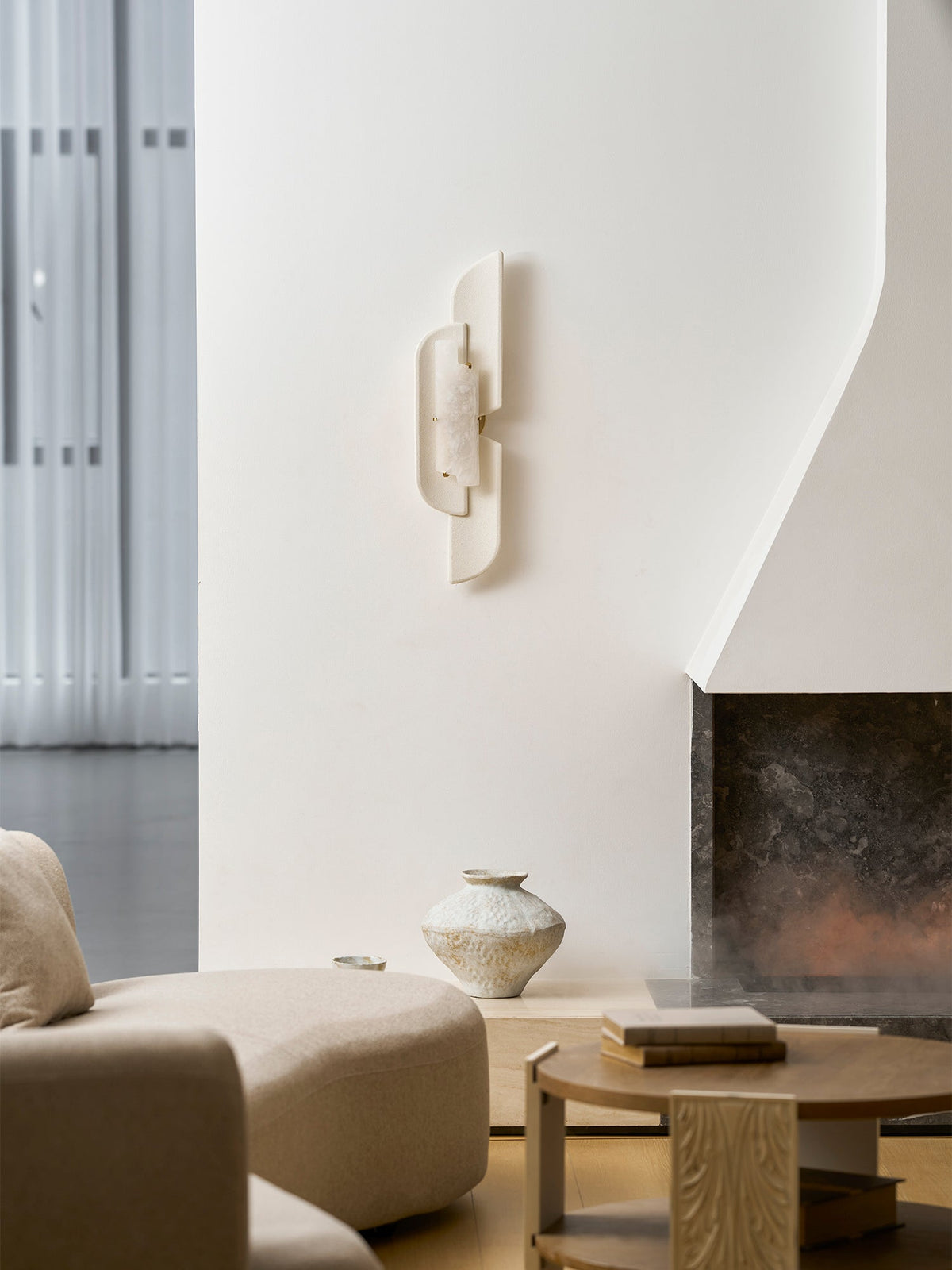 An unlit Clérachic wall lamp on a white plaster wall above a curved sofa and a wooden table, with a fireplace glowing.
