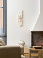 A Clérachic wall lamp on a white wall beside a tall curtain, above a vase, a cream chair and a round table.