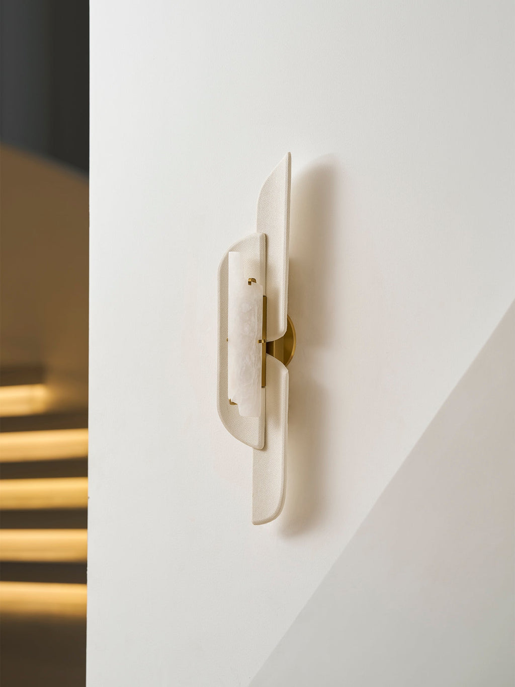 A Clérachic wall lamp with brass fittings shown close to a white wall, with stair lights glowing in the dark behind.