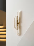A Clérachic wall lamp with brass fittings shown close to a white wall, with stair lights glowing in the dark behind.