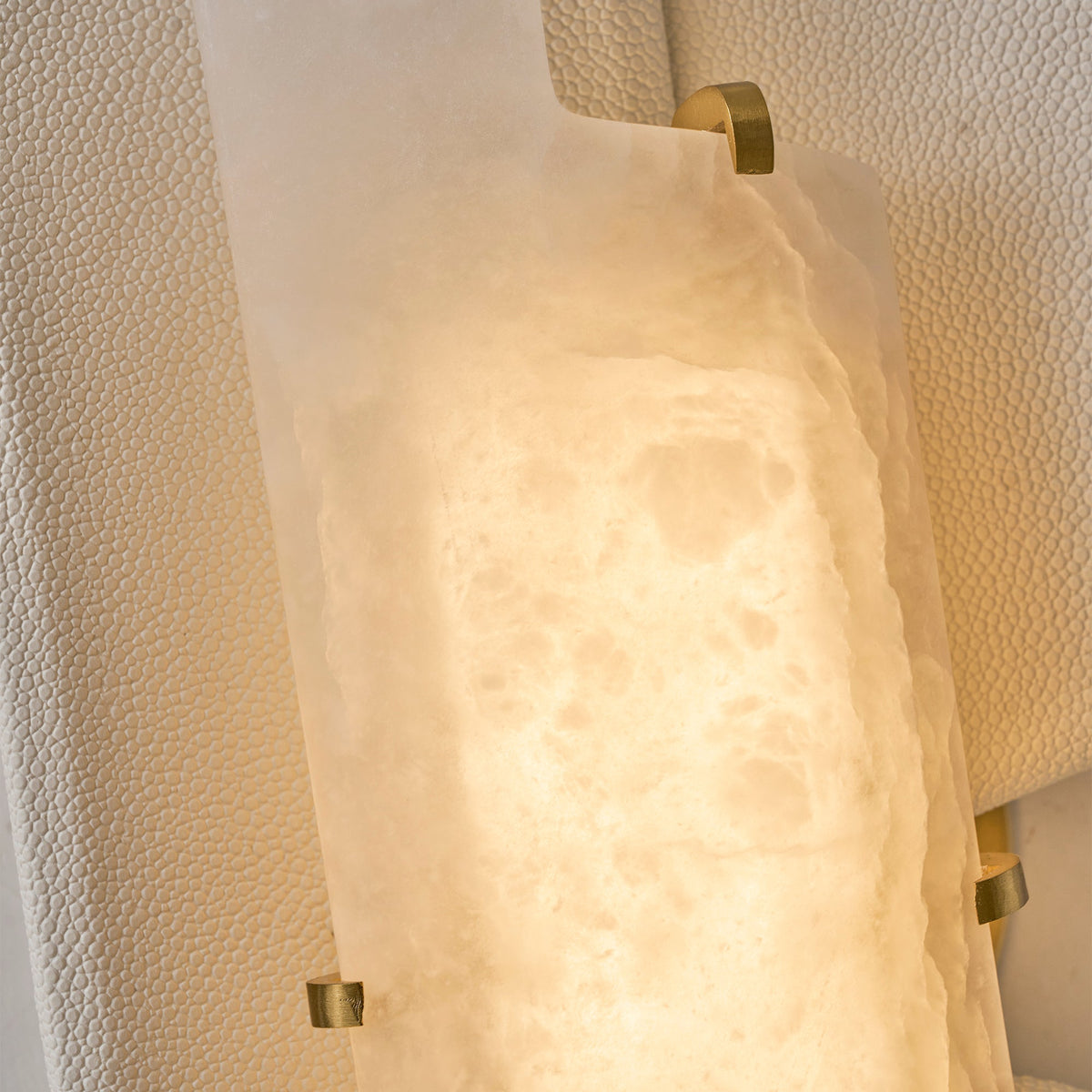 Close view of the lit alabaster slab on this Clérachic wall lamp, held in place by small brass clips on a pebbled panel.