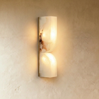 Clérachic double-curved alabaster wall light glowing warm, its veining lit, on a slim gold backplate against plaster.