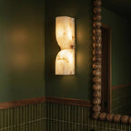 Clérachic alabaster wall light glowing beside a beaded wooden mirror in a green tiled room, veined stone lit from within.