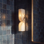 Close view of a Clérachic alabaster wall light glowing against dark blue glazed tiles, with a brass mirror frame at right.