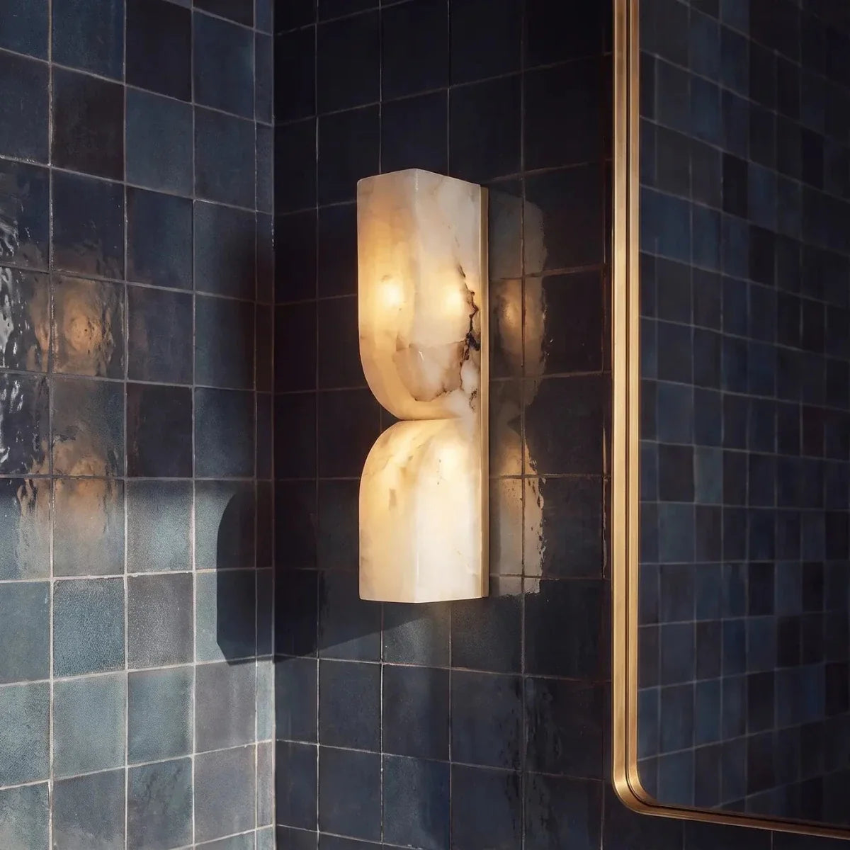 Close view of a Clérachic alabaster wall light glowing against dark blue glazed tiles, with a brass mirror frame at right.