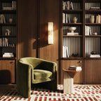 Clérachic tall alabaster wall light on a wood-panelled wall above a green velvet armchair and built-in bookshelves.