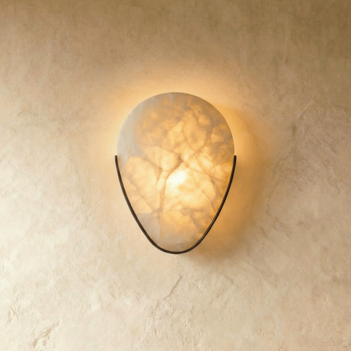 Clérachic alabaster wall sconce glowing warm on a limewashed wall, its veined stone held in a slim dark metal arc.