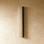 Clérachic tall alabaster bar wall light with a black metal backing, unlit, mounted on a pale limewash wall in a studio shot.