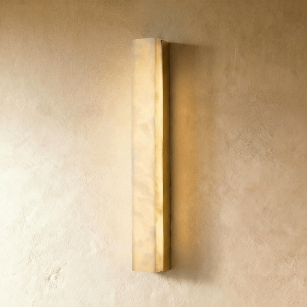 Clérachic medium-length alabaster wall light in a gold frame, casting soft light on a limewash wall in a studio shot.