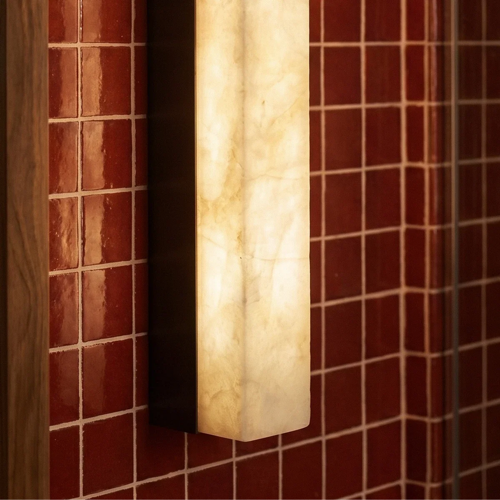 Close up of a Clérachic lit alabaster bar wall light with a black frame against glossy red tiles beside a timber mirror edge.