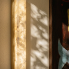 Close up of a Clérachic lit alabaster wall light with a gold edge and veined stone glow, with dappled shadows on the wall.