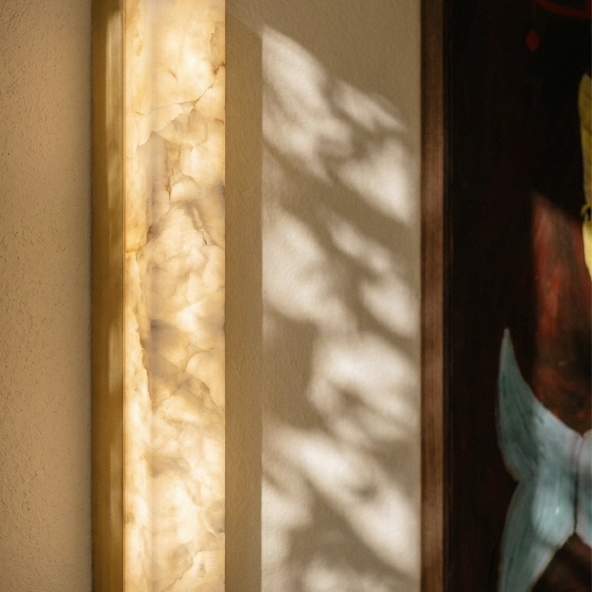Close up of a Clérachic lit alabaster wall light with a gold edge and veined stone glow, with dappled shadows on the wall.