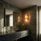 Clérachic two-light alabaster wall lamp glowing beside a brass-framed mirror in a bathroom with leafy mural walls.