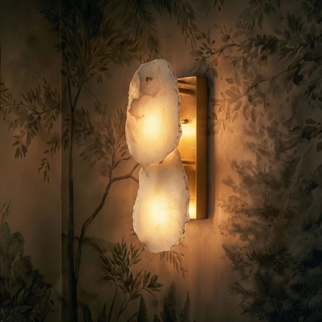 Clérachic two-light alabaster wall lamp lit warm on a brass plate against a soft botanical mural wall.