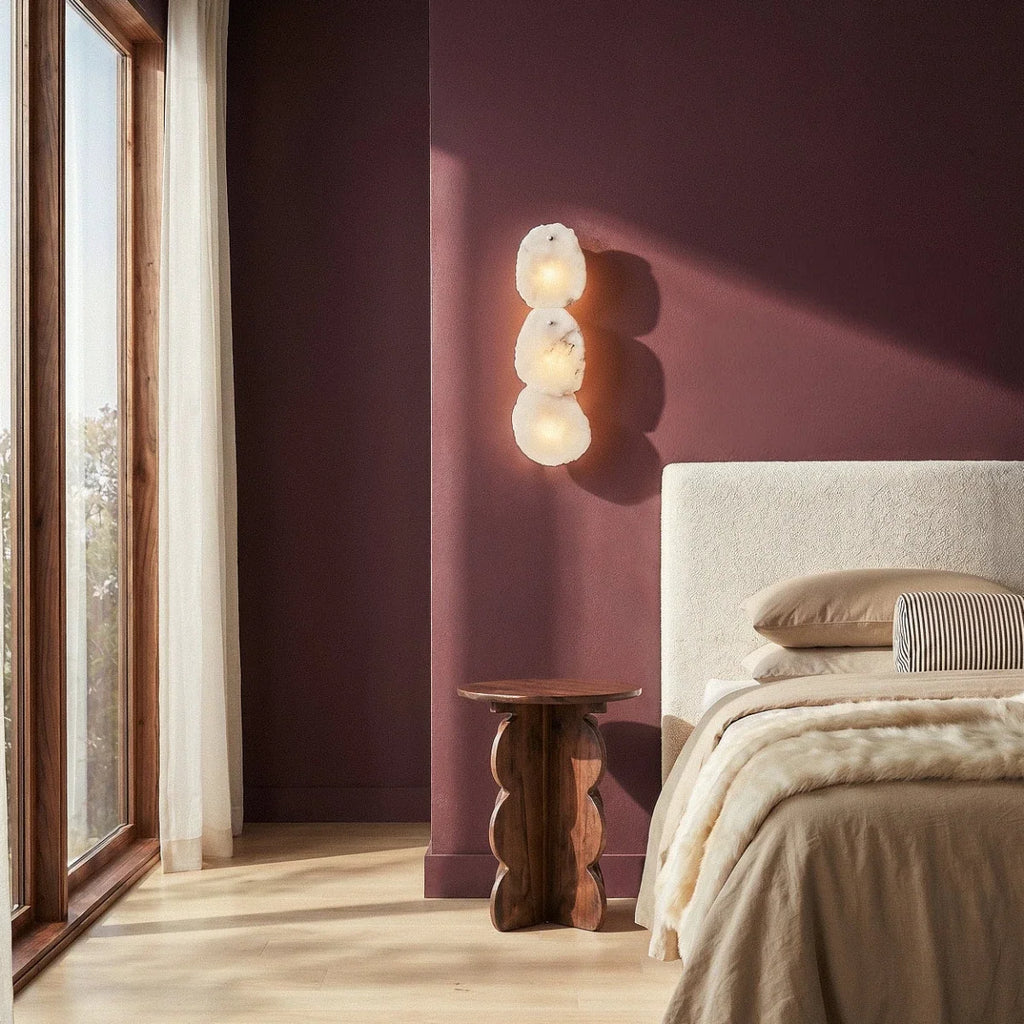 Clérachic three-light alabaster wall lamp above a wooden side table in a plum bedroom beside a window.