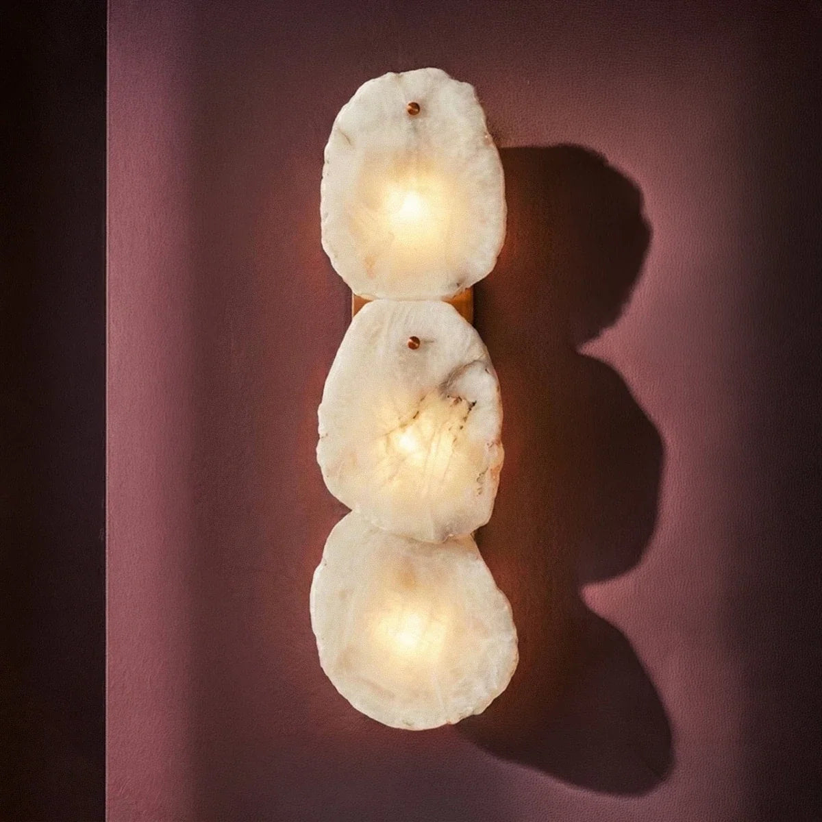 Clérachic three-light alabaster wall lamp glowing on a plum wall, casting soft rounded shadows.