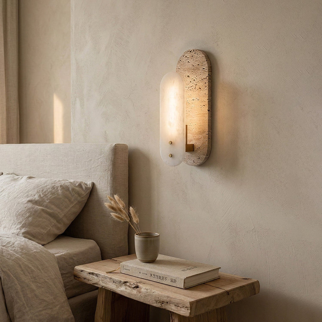 A lit Clérachic wall light glows on a plaster wall above a rough wooden bedside table with a book and a dried-grass vase, beside a linen bed.