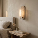 A lit Clérachic wall light glows on a plaster wall above a rough wooden bedside table with a book and a dried-grass vase, beside a linen bed.