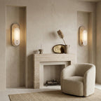 Two Clérachic wall lights glow either side of a plaster fireplace, with a cream bouclé armchair, a dried plume and a brass tray between them.
