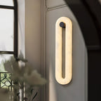 Clérachic Alabaster Wall Sconce lit beside an arched window, glowing softly on a panelled wall above a vase of foliage.