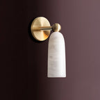 Cylindrical alabaster shade with soft grey veining hanging from the brass ball joint of this Clérachic wall light, on a muted aubergine wall.