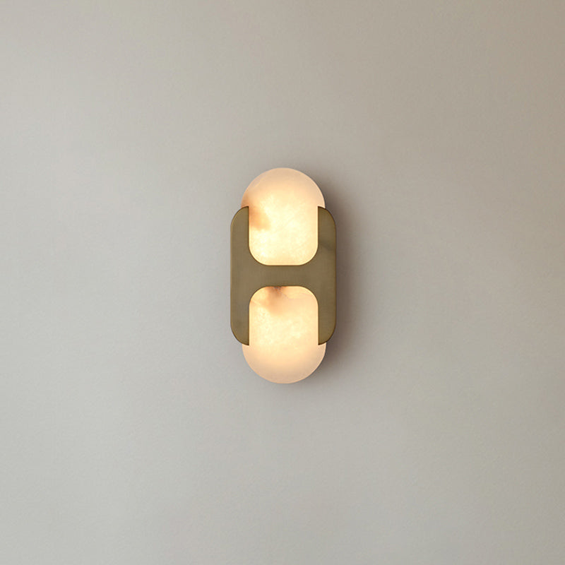 Clérachic Alabaster Wall Sconce in brass, lit on a pale grey wall, its H-bracket splitting the stone into two soft glowing lobes.