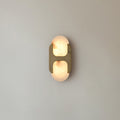 The short Clérachic Alabaster Wall Sconce, brass bracket over chalky white stone, glowing quietly against a plain grey-blue wall.