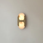 The short Clérachic Alabaster Wall Sconce, brass bracket over chalky white stone, glowing quietly against a plain grey-blue wall.