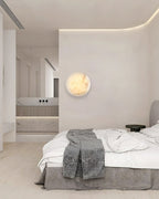 Clérachic Alabaster Wall Sconce lighting a pale bedroom wall beside a made bed, with the bathroom visible beyond.