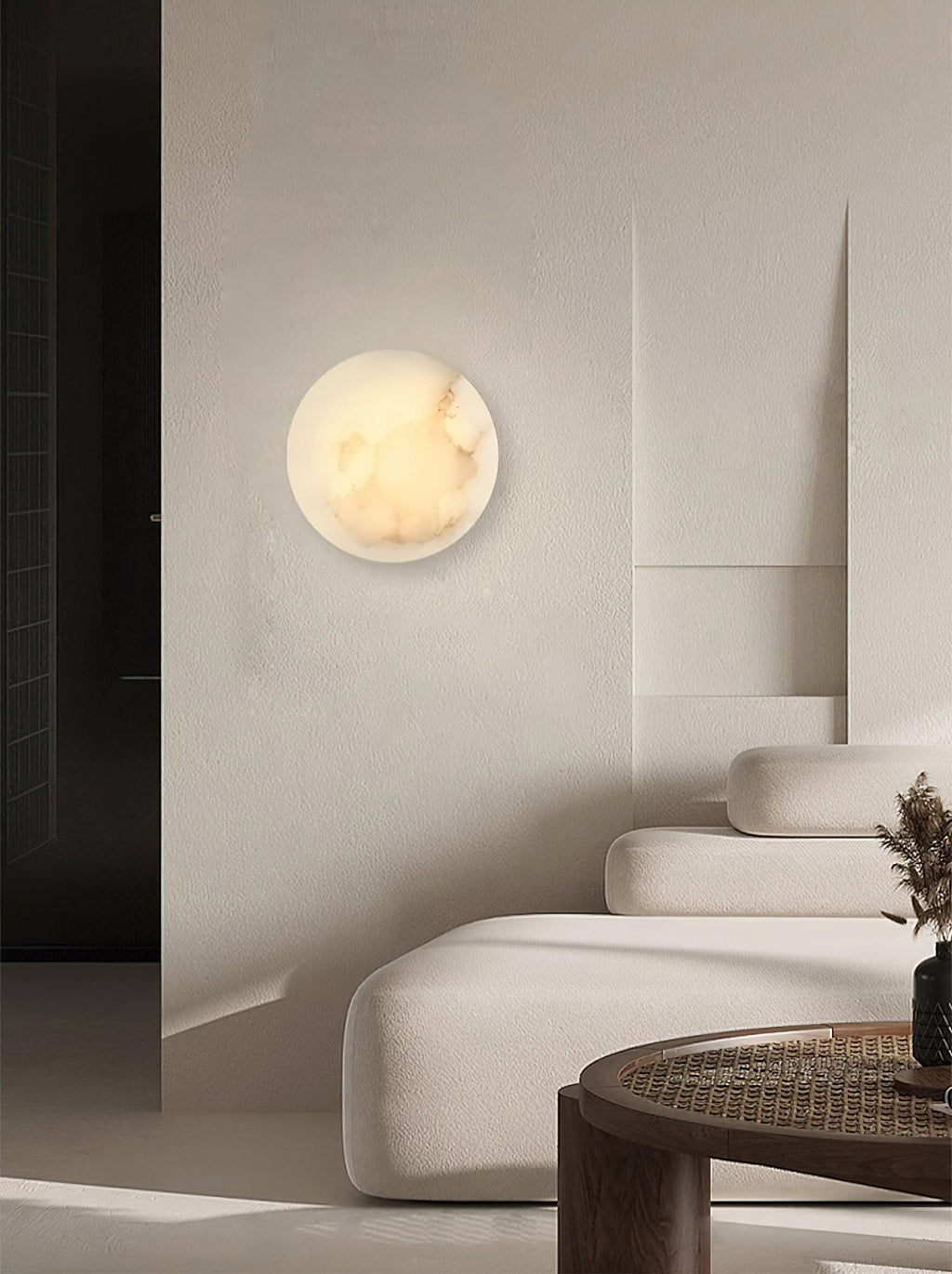 A Clérachic Alabaster Wall Sconce glowing on a plaster living-room wall above a curved boucle sofa and rattan table.