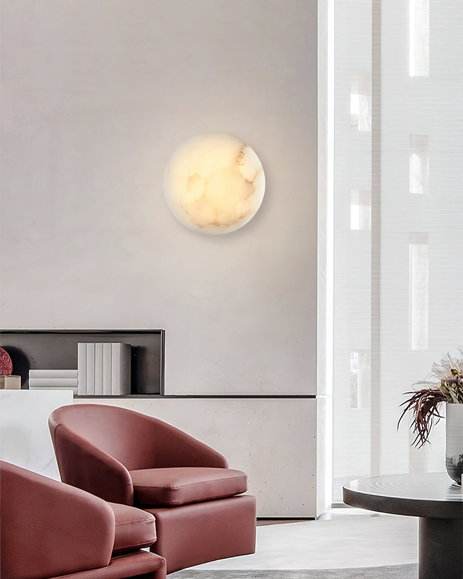 Clérachic Alabaster Wall Sconce glowing on a concrete lounge wall above two oxblood leather armchairs.