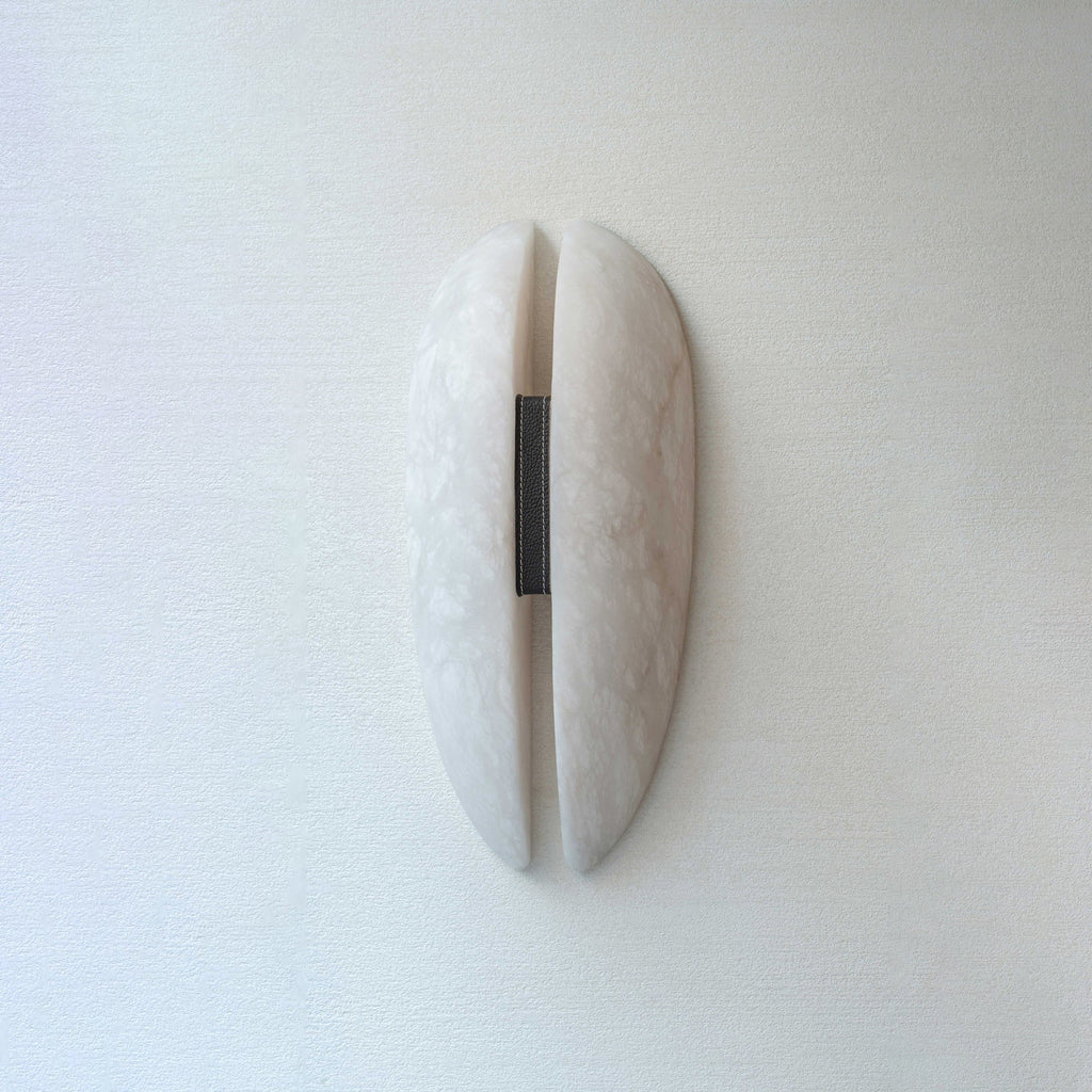Clérachic alabaster wall sconce unlit on a soft white wall, two curved stone panels parted by a slim gap and bound with a leather strap.