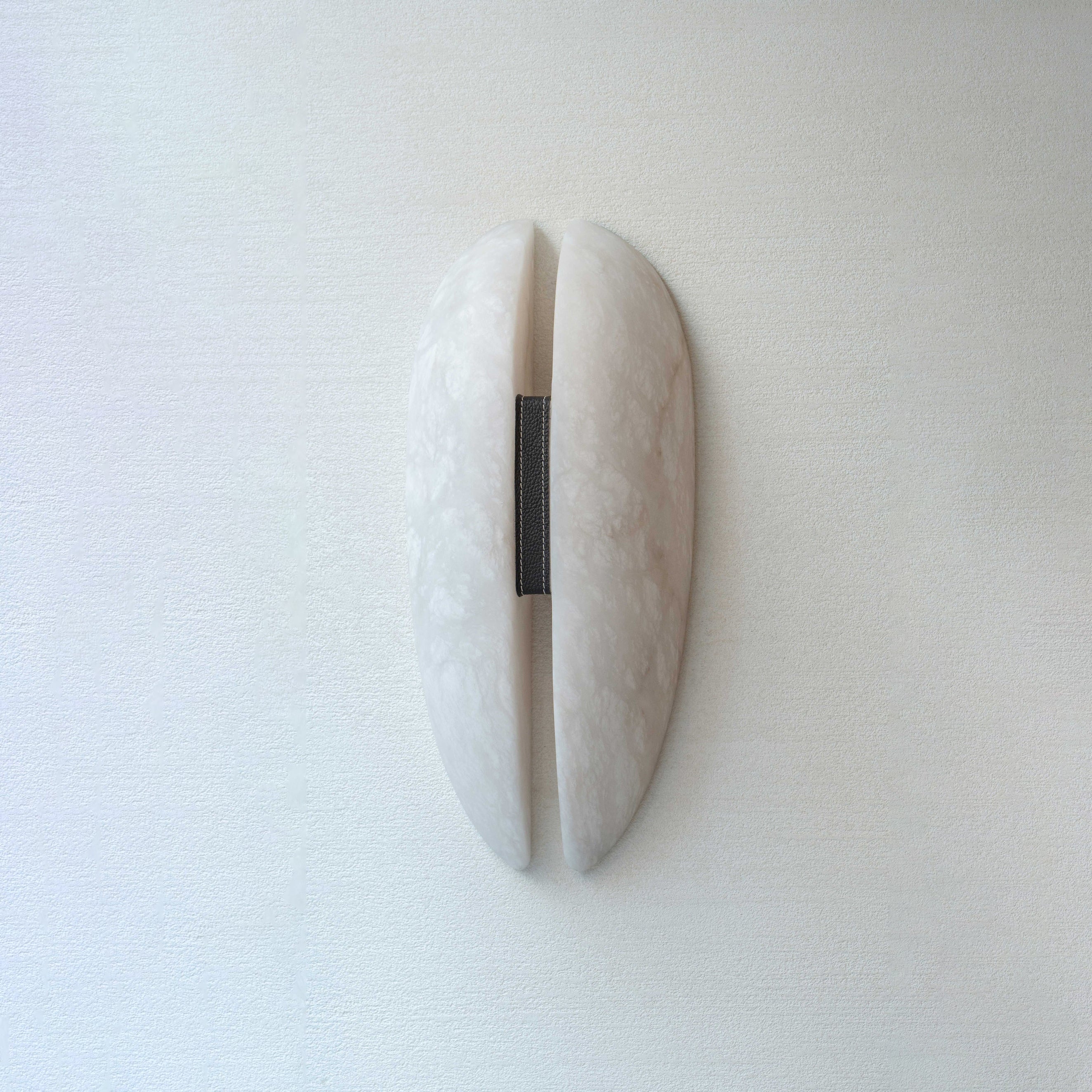 Clérachic alabaster wall sconce unlit on a soft white wall, two curved stone panels parted by a slim gap and bound with a leather strap.