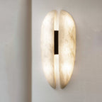 The Clérachic alabaster sconce lit on a pale wall, the slot throwing a warm line up and down while the black leather strap stays dark.