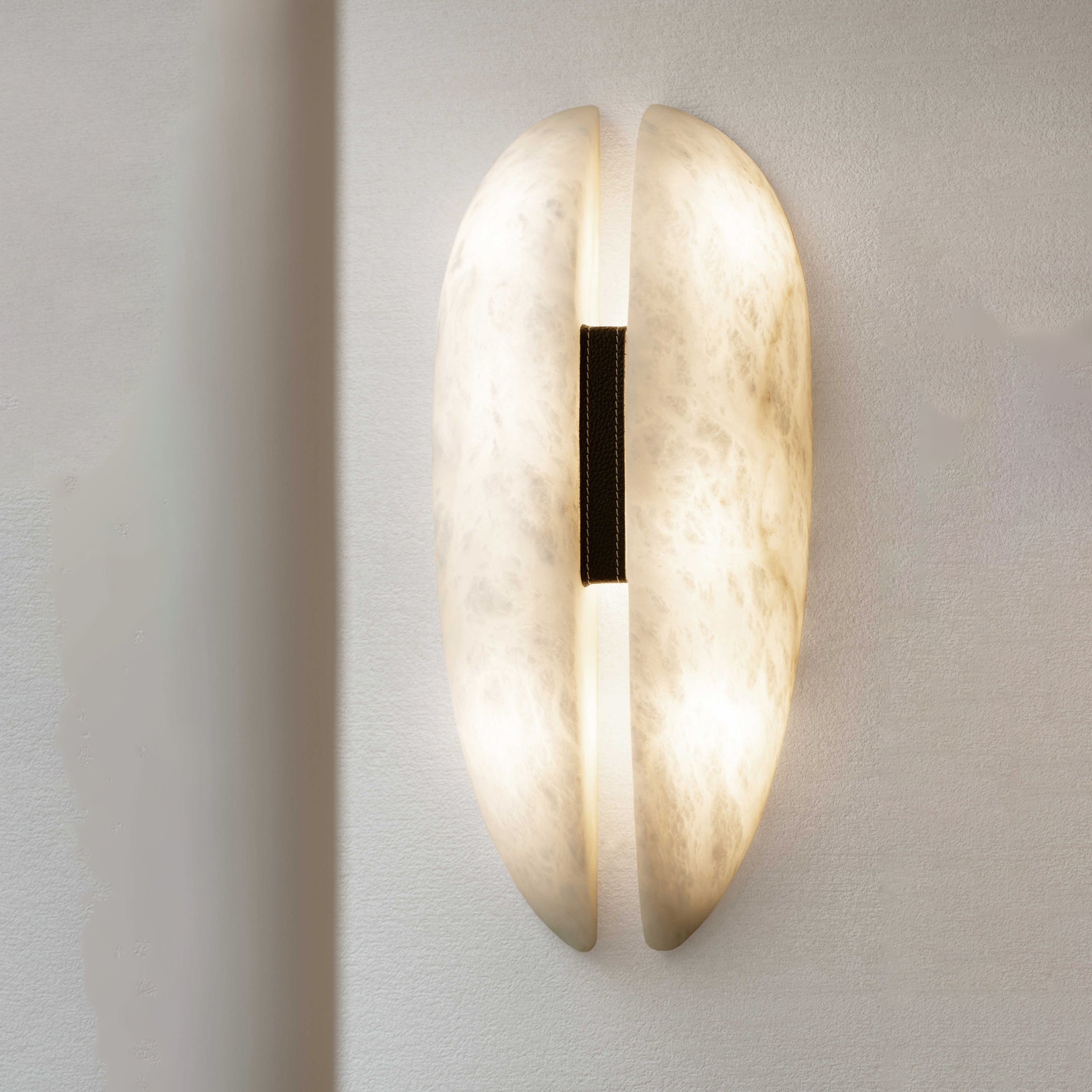 The Clérachic alabaster sconce lit on a pale wall, the slot throwing a warm line up and down while the black leather strap stays dark.