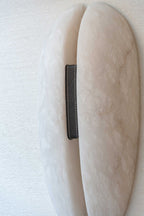 Close-up on the Clérachic sconce's black grain-leather strap and cream saddle stitching where it binds the two alabaster halves.
