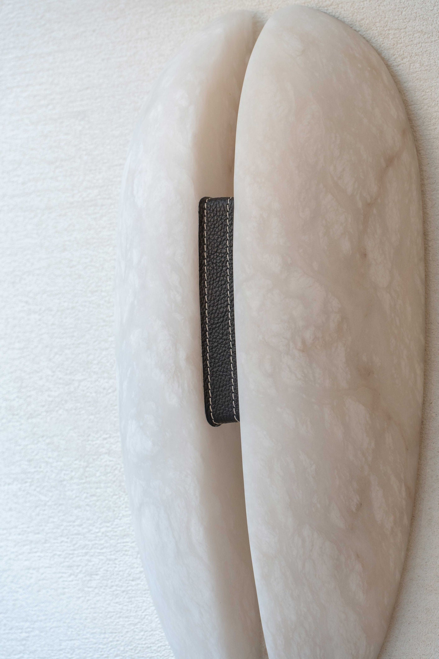 Close-up on the Clérachic sconce's black grain-leather strap and cream saddle stitching where it binds the two alabaster halves.