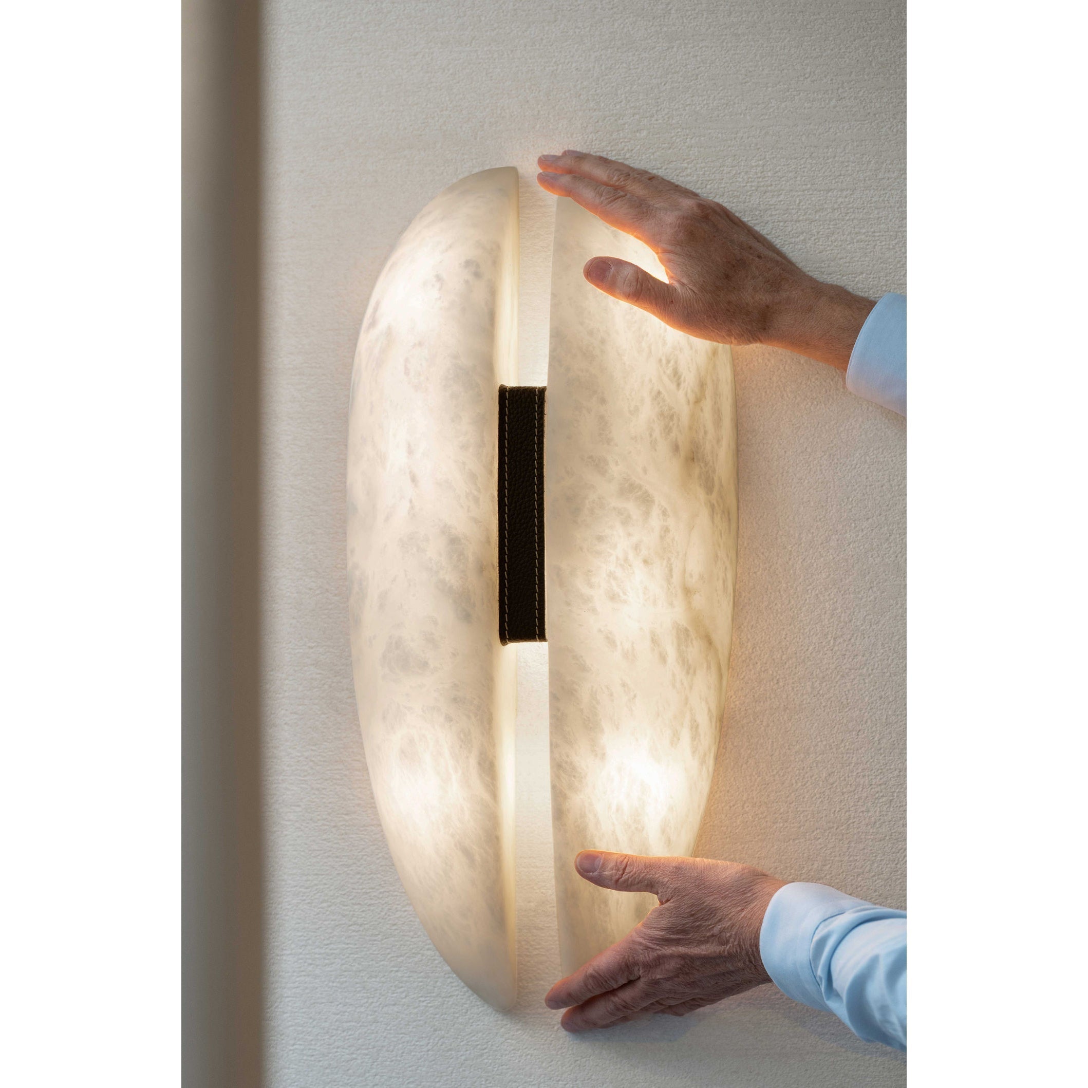 Two hands held either side of the lit Clérachic alabaster sconce, showing how the larger size fills a stretch of wall.