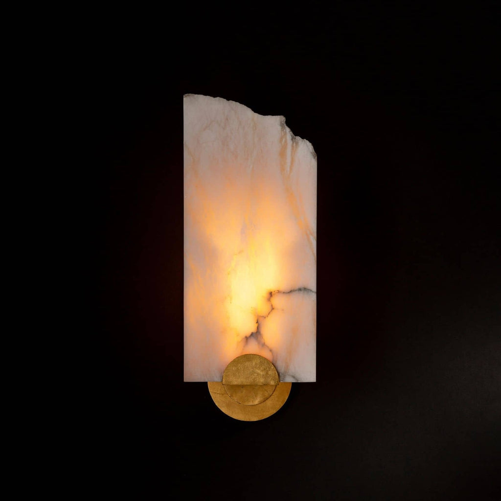Clérachic Alabaster Wall Sconce glowing in a darkened room, the stone veining lit from within above a round brass base.