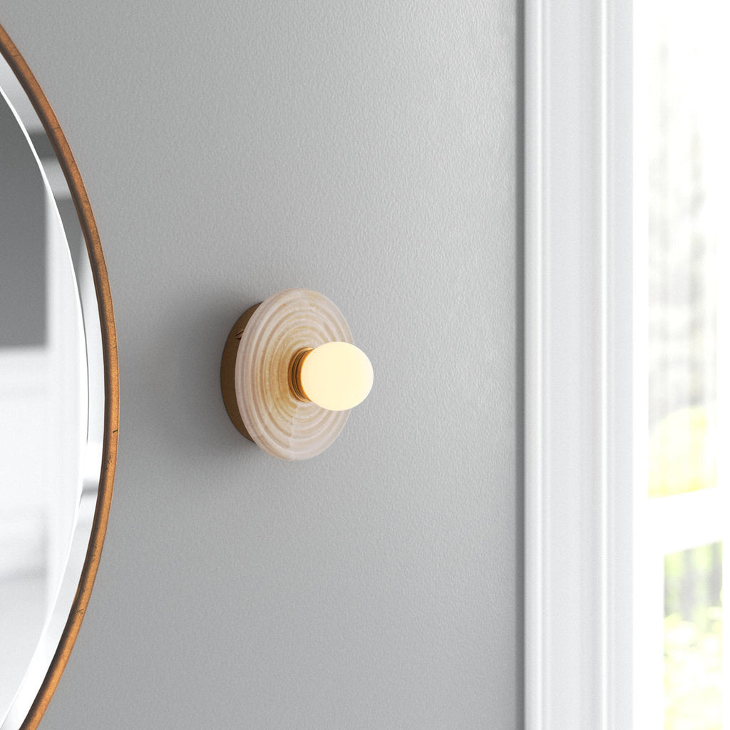 Gold Clérachic alabaster sconce lit on a soft grey wall beside a round brass mirror, sitting close to the wall in a bright corner.