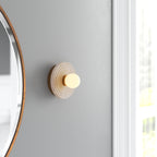 Gold Clérachic alabaster sconce lit on a soft grey wall beside a round brass mirror, sitting close to the wall in a bright corner.