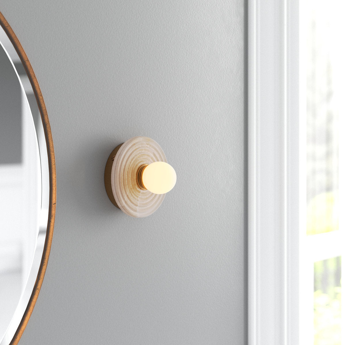 Gold Clérachic alabaster sconce lit on a soft grey wall beside a round brass mirror, sitting close to the wall in a bright corner.