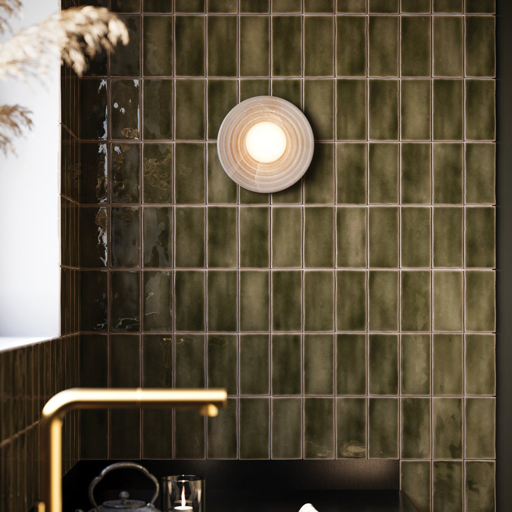Clérachic alabaster sconce lit against a deep green tiled wall above a counter, its warm glow picking out the glaze of each tile.