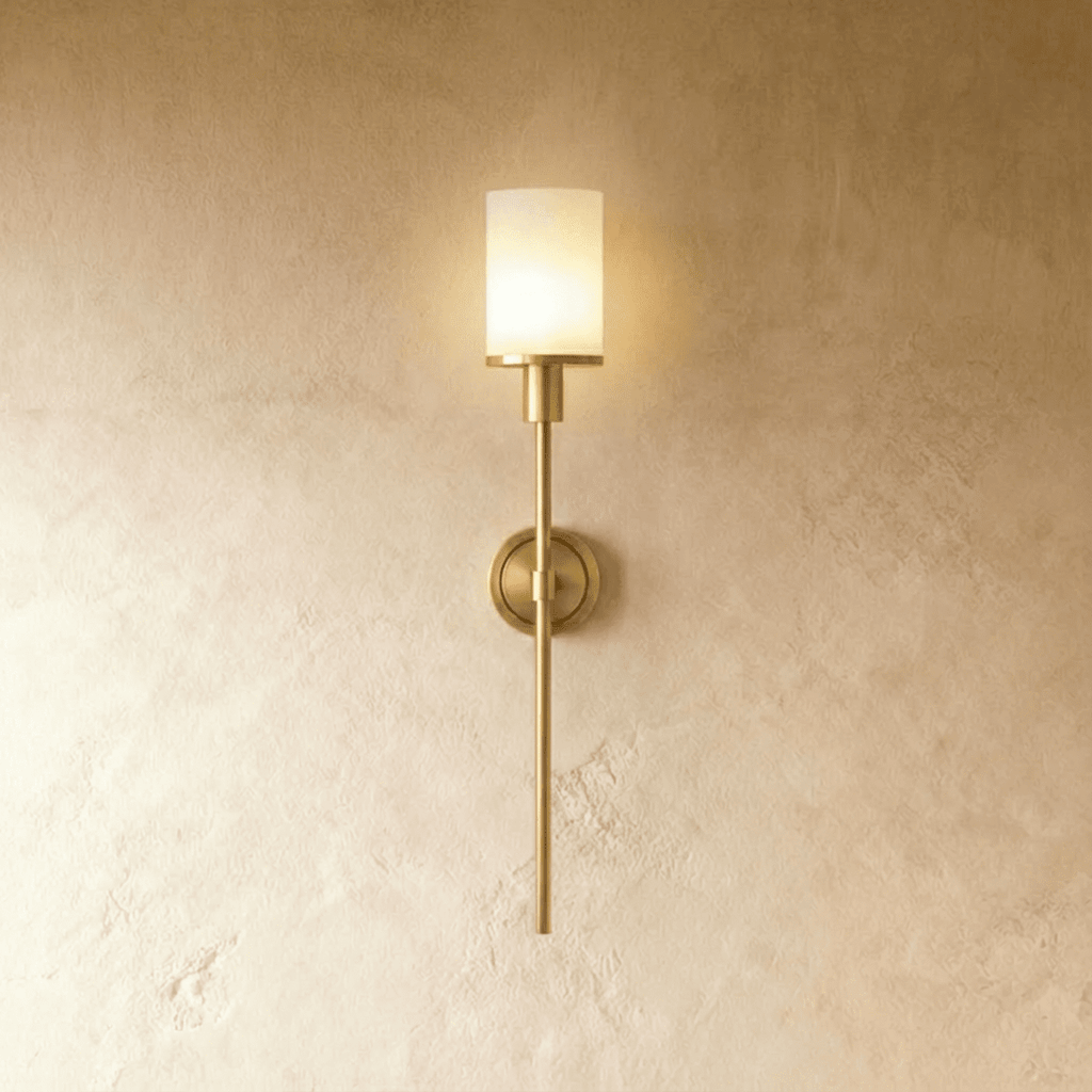 Clérachic brass wall sconce with a glowing white cylinder shade, round backplate and slim rod on a limewash wall.