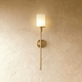 Clérachic brass wall sconce with a glowing white cylinder shade, round backplate and slim rod on a limewash wall.