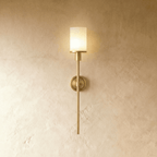 Clérachic brass wall sconce with a glowing white cylinder shade, round backplate and slim rod on a limewash wall.